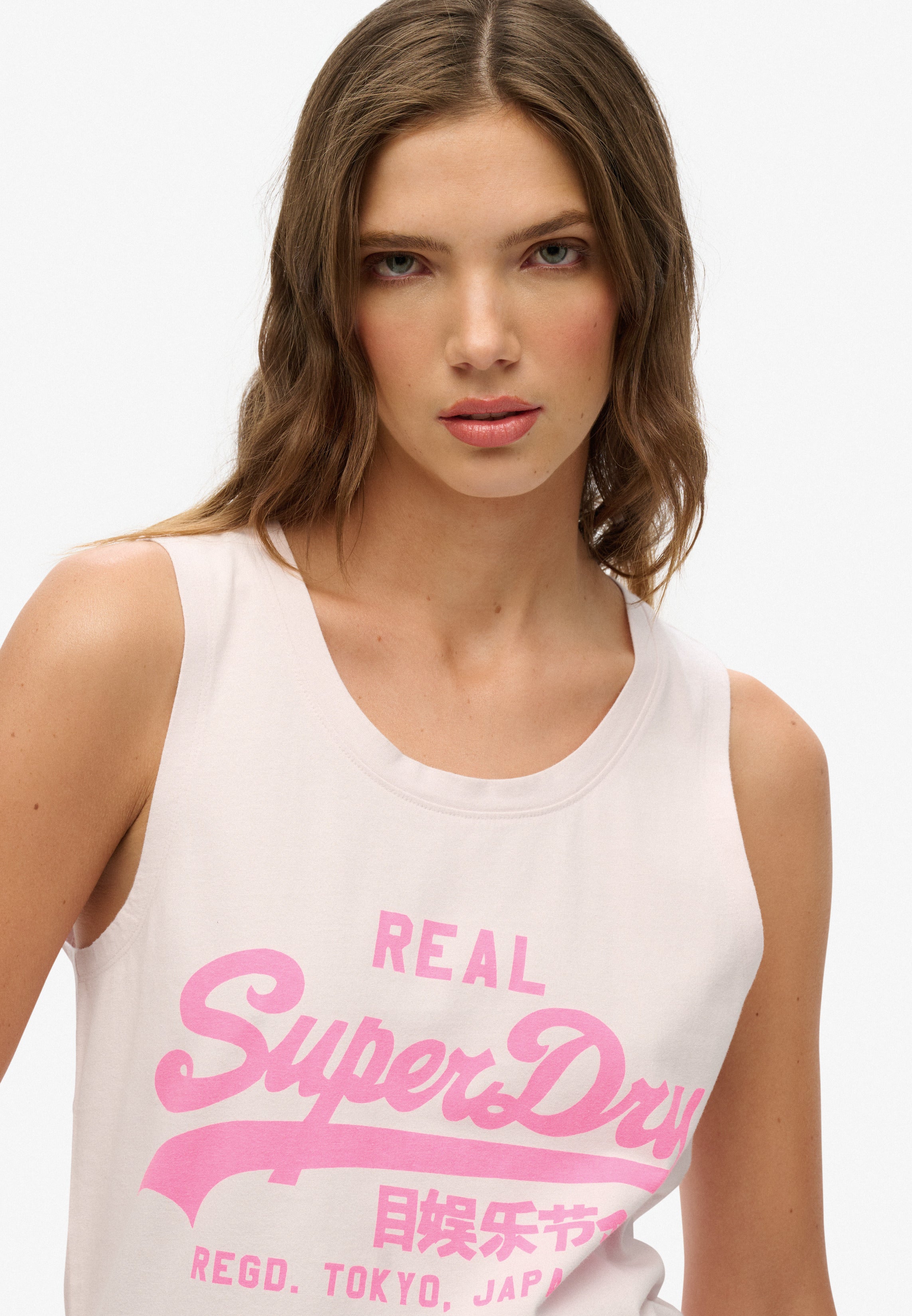 Woman wearing a white tank top with pink 'Superdry' logo on a white background